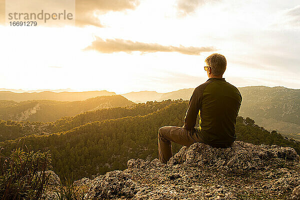 Mann betrachtet den Sonnenuntergang auf einem Felsen sitzend vom Gipfel eines Berges aus