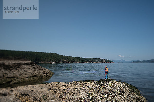 Junges Mädchen  das auf einem Felsen steht und unter blauem Himmel auf das Meer hinausschaut  auf Lopez Island WA