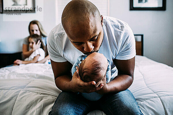 Vater küsst Neugeborenes auf Bett mit Mutter und Tochter im Hintergrund