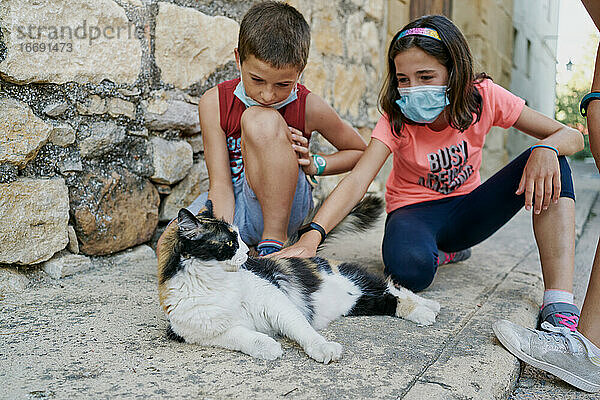 Zwei Kinder mit Masken für das covid19 spielen mit einer schwarz-weißen Katze  die auf der Straße liegt