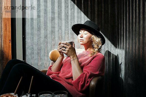 Frau sitzt mit einem warmen Getränk in der Hand in einem Café und genießt die Sonne