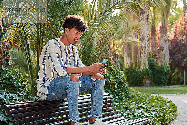 Ein junger Mann mit Afro-Haar  der auf einer Bank sitzend sein Smartphone beobachtet