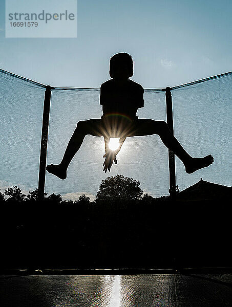 Vertikale Hintergrundbeleuchtung Foto einer Silhouette eines barfuß springenden oder fliegenden Jungen auf einem Trampolin mit Netz und die Sonne zwischen den Armen reflektiert Sonnenstrahlen  die einen Regenbogen erzeugen