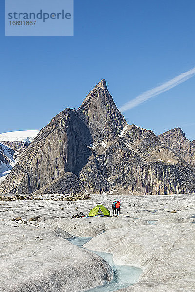 Zwei Bergsteiger stehen neben einem Zelt auf einem Gletscher unter einem Berggipfel.