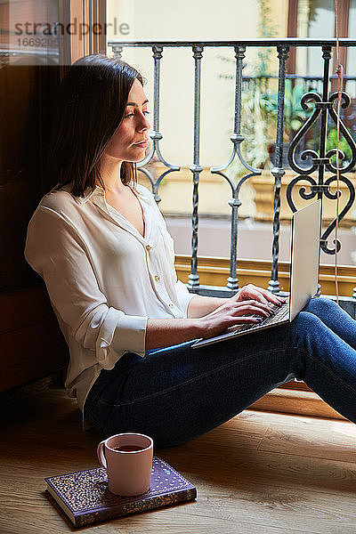 Junge Frau arbeitet von zu Hause aus mit einem Laptop neben einem Fenster.
