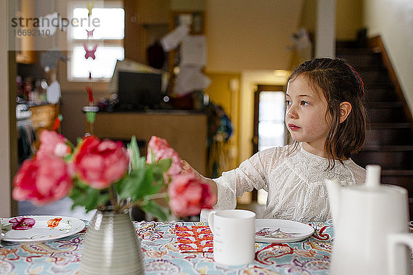 Ein kleines Kind in einem Spitzenkleid sitzt allein an einem für eine Teeparty gedeckten Tisch