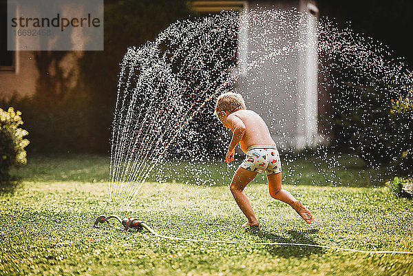 Junge weiße Junge läuft unter dem Wasser aus dem Sprinkler im Garten