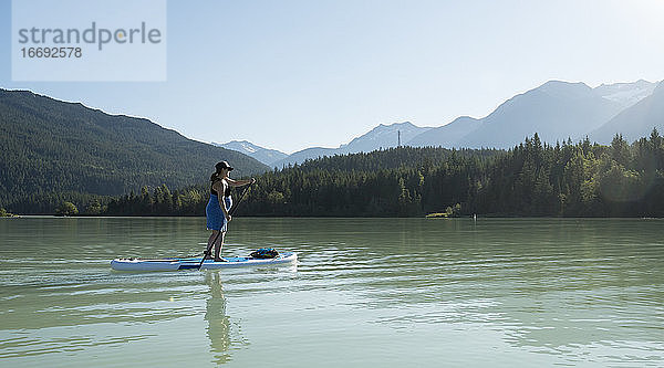 Barfüßige Frau auf einem Paddelbrett auf einem See vor einem grünen Bergkamm an einem sonnigen Sommertag in British Columbia  Kanada