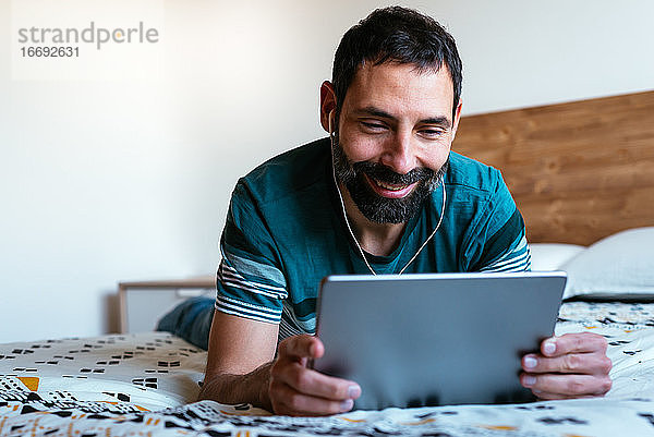 Junger Mann liegt auf dem Bett und schaut auf ein digitales Tablet mit Kopfhörern