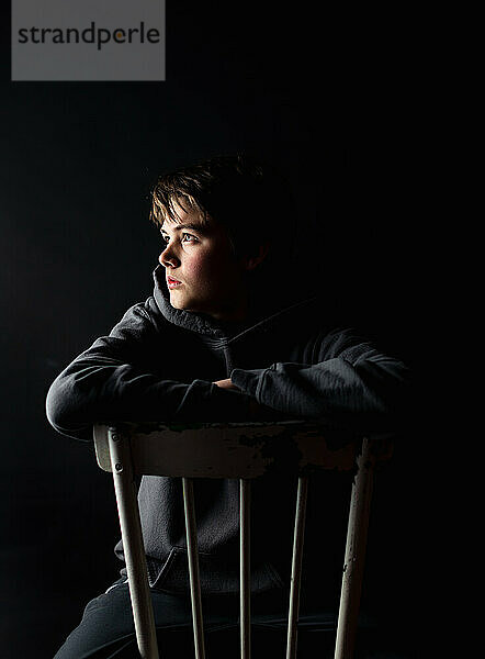 Low-Key-Porträt eines heranwachsenden Jungen  der auf einem Stuhl in einem dunklen Raum sitzt.