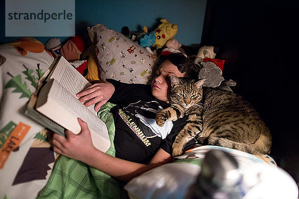 Zehnjähriger Junge liest im Bett ein Buch  während eine getigerte Katze sein Gesicht kuschelt