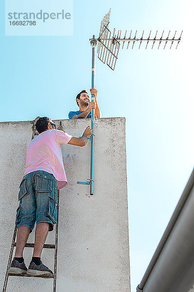 Zwei Männer installieren an einem sonnigen Tag eine Fernsehantenne auf dem Hochdach eines Landhauses.