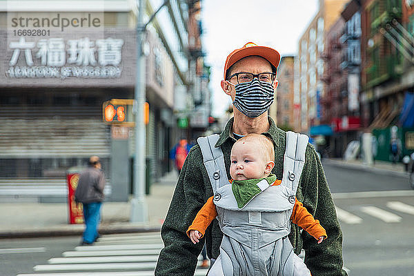 Vater mit Gesichtsmaske  der seine Tochter in der Babytrage trägt  während er in der Stadt während der Pandemie die Straße überquert