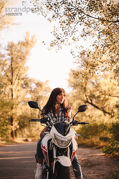 Selbstbewusste junge Frau auf einem Motorrad auf einer Landstraße im Wald sitzend