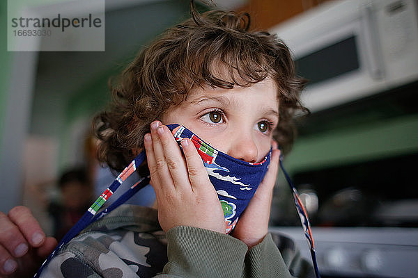 Kleiner Junge  der vor dem Verlassen des Hauses Hilfe beim Aufsetzen seiner Maske erhält