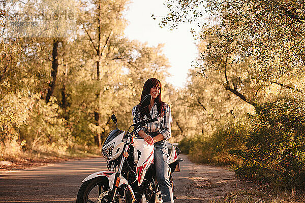 Lächelnde junge Frau auf Motorrad sitzend auf Landstraße inmitten von Bäumen
