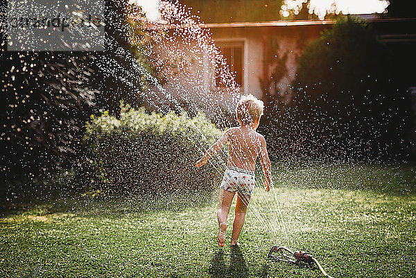 Kleines Kind läuft unter dem Wasser aus dem Sprinkler im Garten