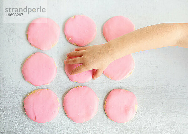 Draufsicht Kinderhand  die rosa Zuckerplätzchen vom Tisch nimmt