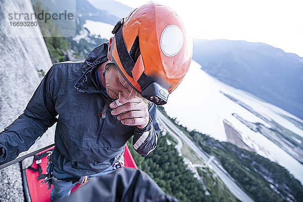 Bergsteiger mit Helm  der sich an der Nase kratzt  steht auf dem Portaledge bei Sonnenuntergang