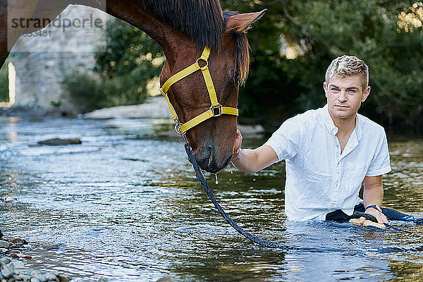 Porträt eines blonden jungen Mannes mit einem Pferd an einem Fluss