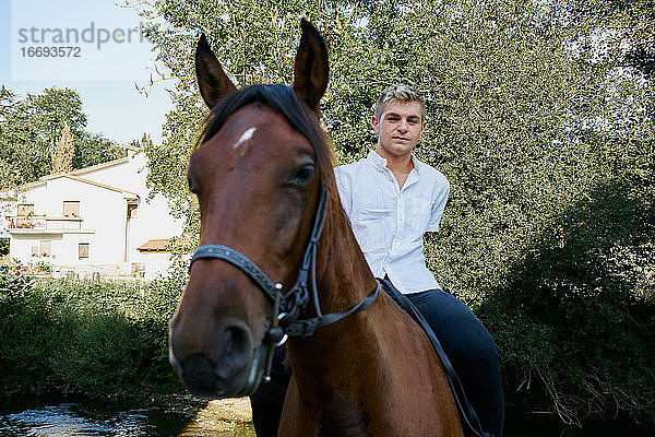 Porträt eines jungen blonden Mannes  der auf einem Pferd über einen Fluss reitet