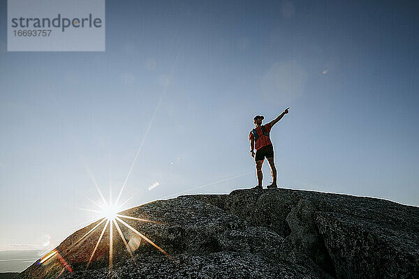 Männlicher Wanderer zeigt auf Aussicht vom felsigen Gipfel eines Berges mit Sonnenstern