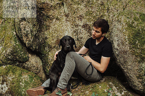 Junger Mann mit schwarzem Labrador Retriever auf einem moosbewachsenen Felsen sitzend