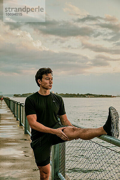 Männlicher Sportler  der sich bei Sonnenuntergang am Ufer dehnt.
