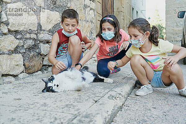 Drei Kinder mit Masken für das covid19 spielen mit einer schwarz-weißen Katze  die auf der Straße liegt