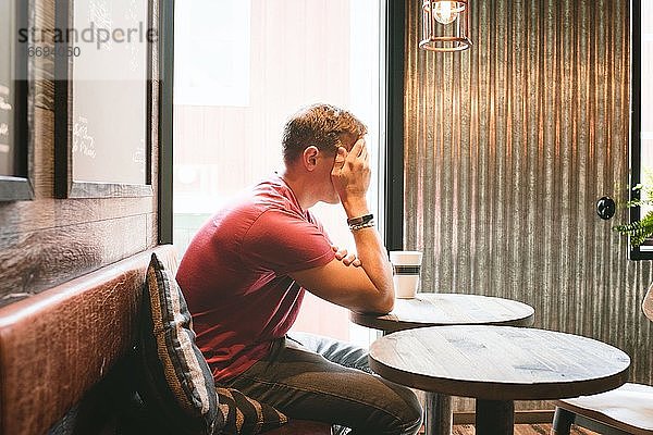 ein Mann  der sein Gesicht bedeckt und gestresst aussieht  während er in einem Cafe sitzt und trinkt