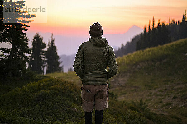 Rückansicht eines Wanderers in einem bauschigen Mantel  der einen farbenfrohen Sonnenuntergang beobachtet