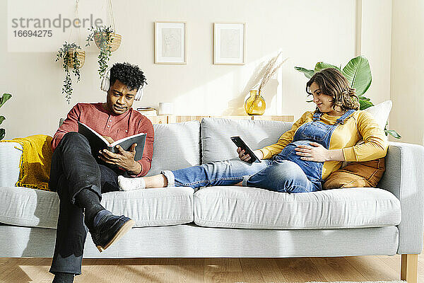 Ehemann hört Musik und liest ein Buch mit seiner schwangeren Frau  die auf dem Sofa ein Tablet betrachtet. Interracial Paar Konzept