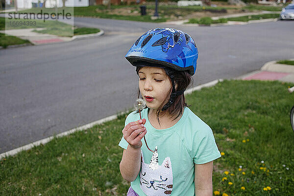 Ein kleines Mädchen mit einem Fahrradhelm pustet die Samen einer Löwenzahnblüte