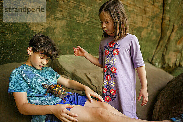 Ein Junge lehnt mit seiner Schwester an einer Felswand und hält eine kleine Schildkröte