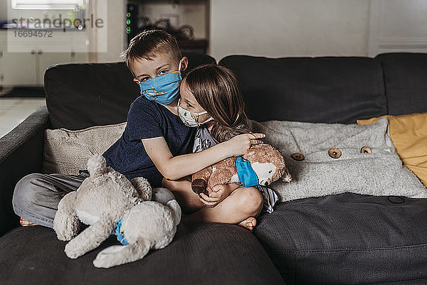 Junges Mädchen und Junge im Schulalter mit Masken  die sich auf der Couch umarmen und lächeln