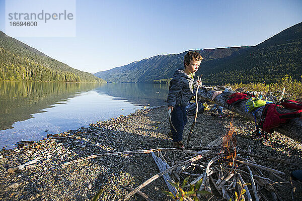 Kleiner Junge trägt Stöcke  um das Feuer am See während eines Campingausflugs zu entzünden