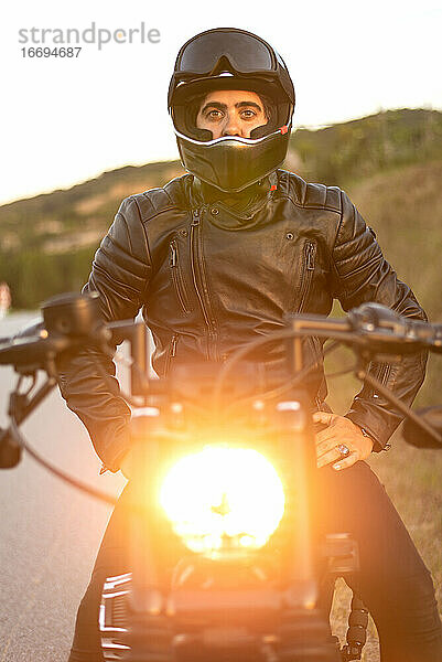Biker mit Helm sitzt und entspannt auf einem alten Motorrad bei Sonnenuntergang.