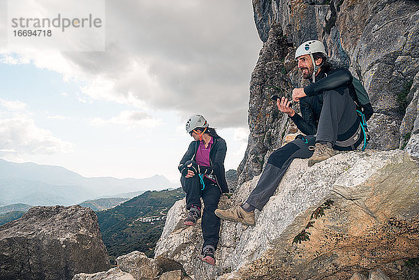 Konzept: Abenteuer. Ein Paar Kletterer mit Helm und Gurtzeug. Sie ruhen sich auf einem Felsen sitzend aus. Sie benutzen das Smartphone und schauen auf den Horizont. Via ferrata in den Bergen.