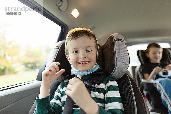 Junge im Grundschulalter lächelt  während er im Auto mit Gesichtsmaske sitzt