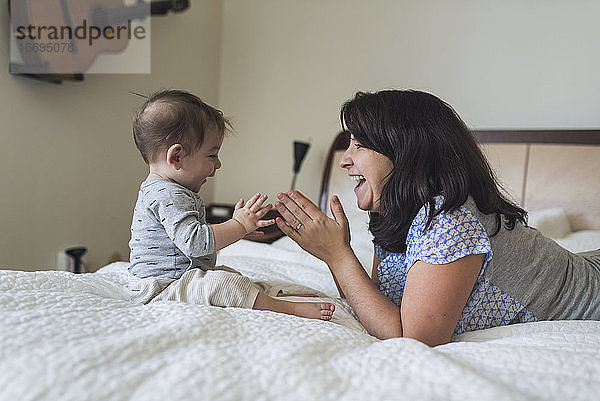 Lachende Mutter Mitte 30  die mit ihrem 1 Jahr alten Baby auf dem Bett in die Hände klatscht