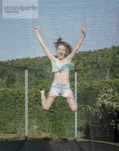 Vertikales Foto eines lächelnden jungen brünetten Teenagers  der auf einem Trampolin mit Netz auf dem grünen Hof im Freien springt. Das Mädchen trägt kurze Jeans und ein Top und sieht Spaß  aktiv