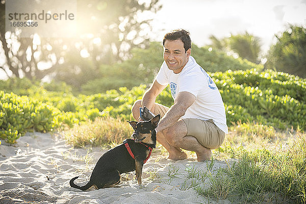 Junger fitter Mann hockt am Strand mit seinem kleinen schwarzen Hund in Hawaii