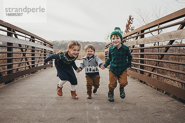 Drei Geschwister halten sich an den Händen und laufen auf einer Brücke in Richtung Kamera