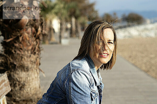 Lächelnde Frau sitzt auf einer Bank in der Nähe des Strandes und schaut in die Kamera