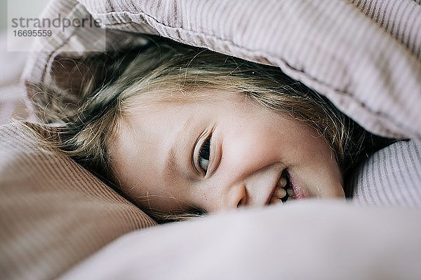 Nahaufnahme offenes Porträt von Mädchen im Bett lächelnd versteckt in den Decken