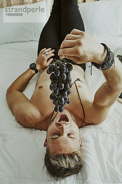 Ein junger Mann liegt im Bett auf dem Rücken und isst eine Traube schwarzer Weintrauben