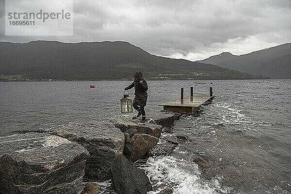 Eine Frau trägt eine Laterne entlang eines steinernen Stegs an einem Fjord