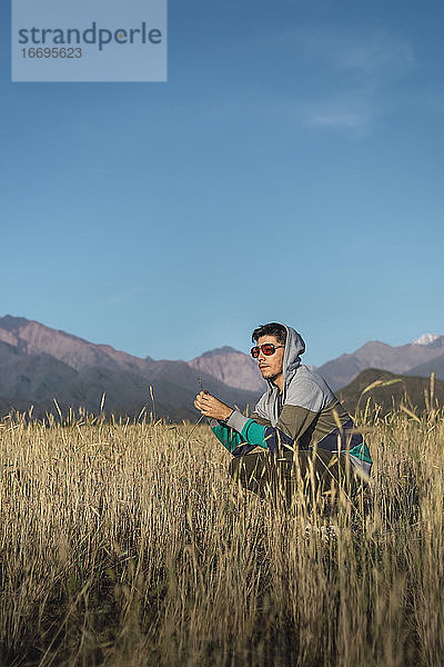 Junger Mann mit Sonnenbrille auf einem Feld sitzend und mit Blick auf die Berge