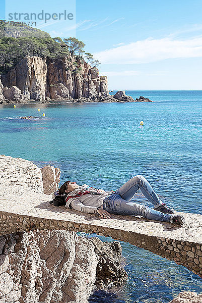 Rückansicht einer Frau  die sich auf einer Brücke liegend vor dem blauen Meer entspannt