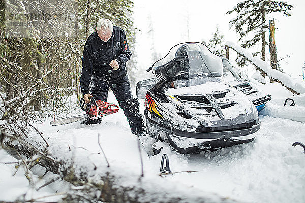 Ein Mann im Ruhestand benutzt eine Kettensäge  um beim Motorschlittenfahren Wege zu räumen.